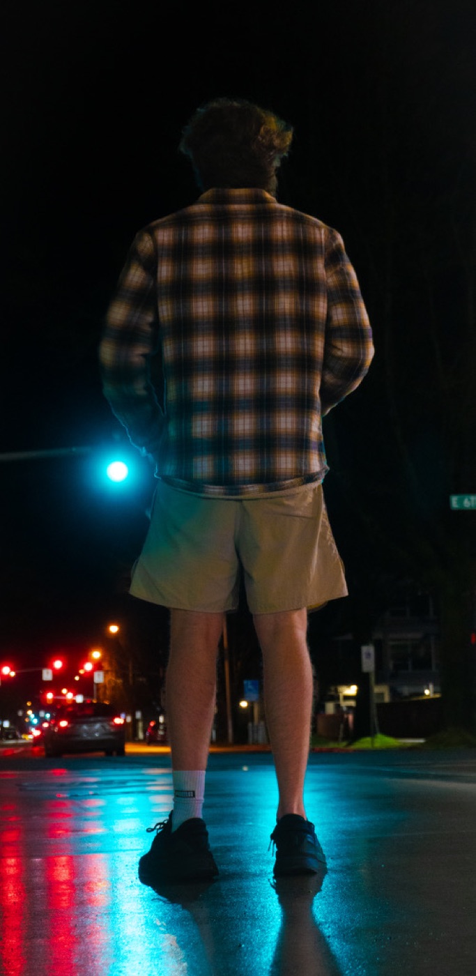 Finn from behind crossing a rain-slick street at night, traffic lights reflected in the road